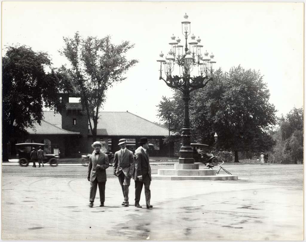 Three men standing near the entrance to Forest Park at Kingshighway Boulevard, with the massive Gilded Age candelabra lamp visible on its raised stone platform in the background. Early automobiles and a horse-drawn carriage are also visible.